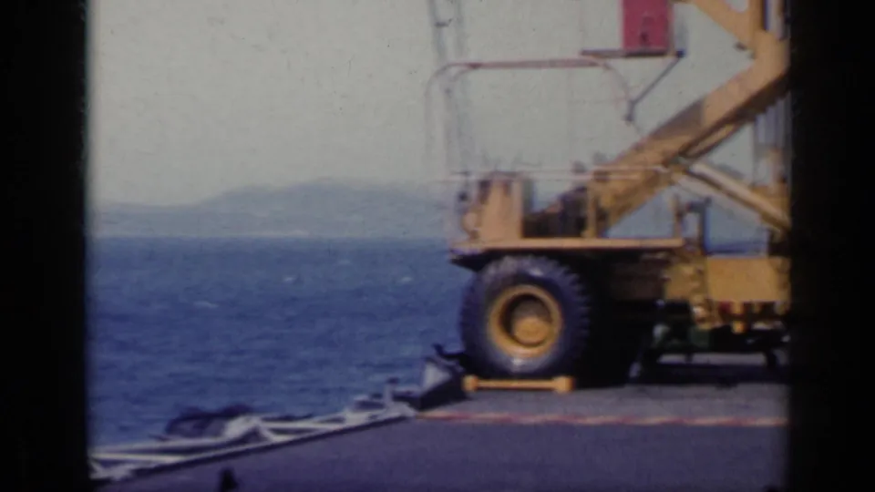 1962 Italy 8mm frame showing a yellow dock crane, pier edge, blue sea and faint coastline in the distance.