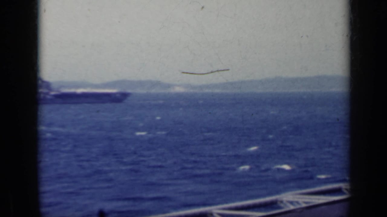 Distant aircraft carrier at sea with mountains on the horizon and dark film borders.