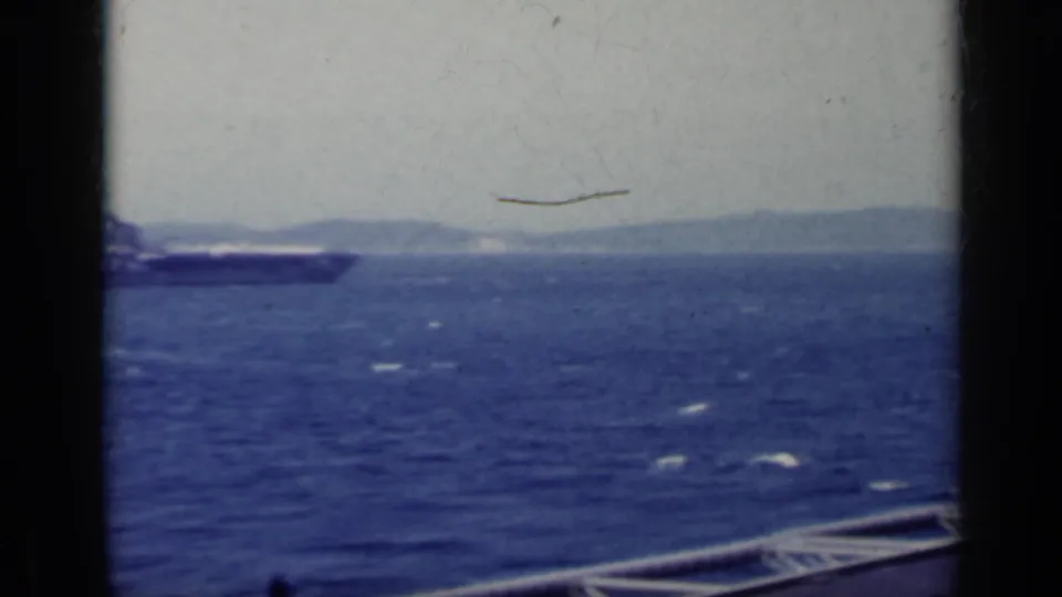 1962 Italy 8mm frame showing a distant aircraft carrier on blue water with hazy mountains beyond.