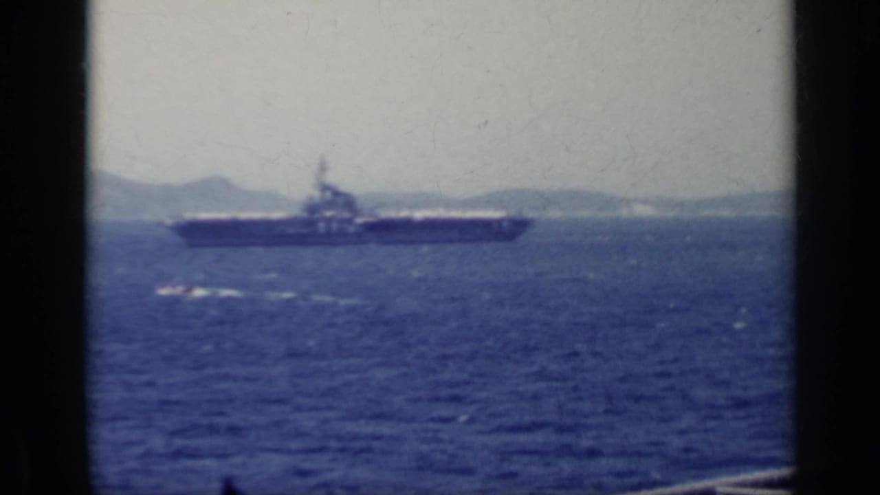 A 1962 Italy home-movie frame shows an aircraft carrier crossing blue water below a pale sky.