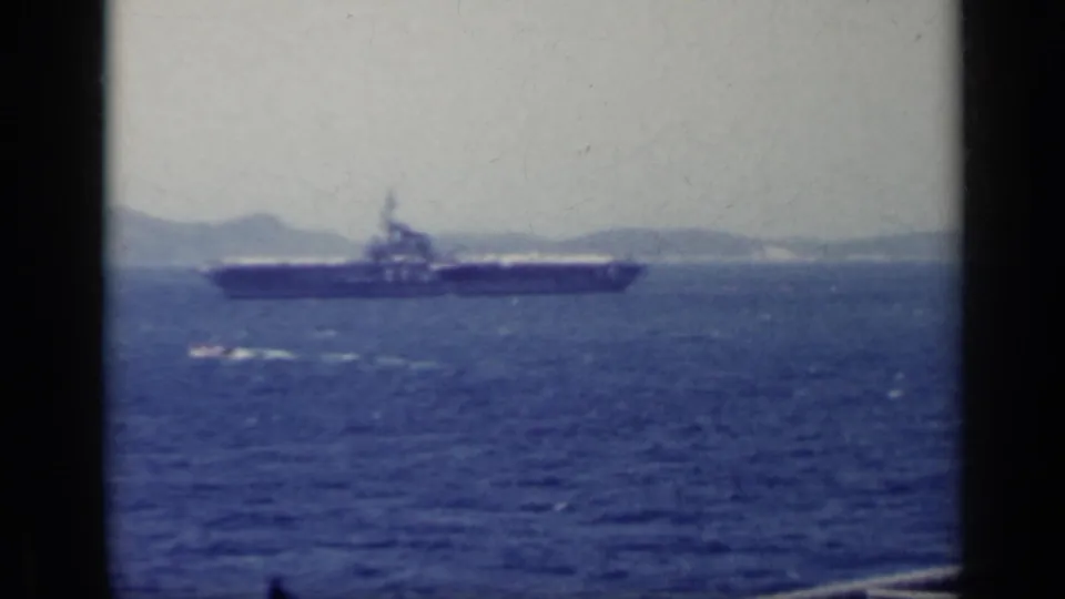 A 1962 Italy home-movie frame shows an aircraft carrier crossing blue water below a pale sky.