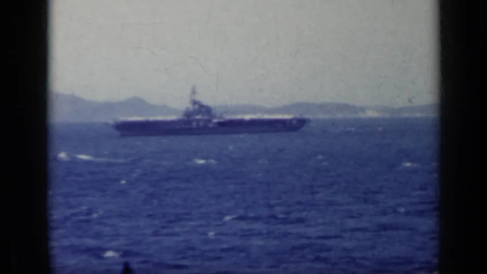 Soft 8mm view of an aircraft carrier offshore in Italy, with blue water and hazy mountains visible.