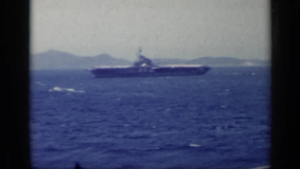 1962 Italy 8mm still of a distant aircraft carrier on blue sea against a low mountain horizon.