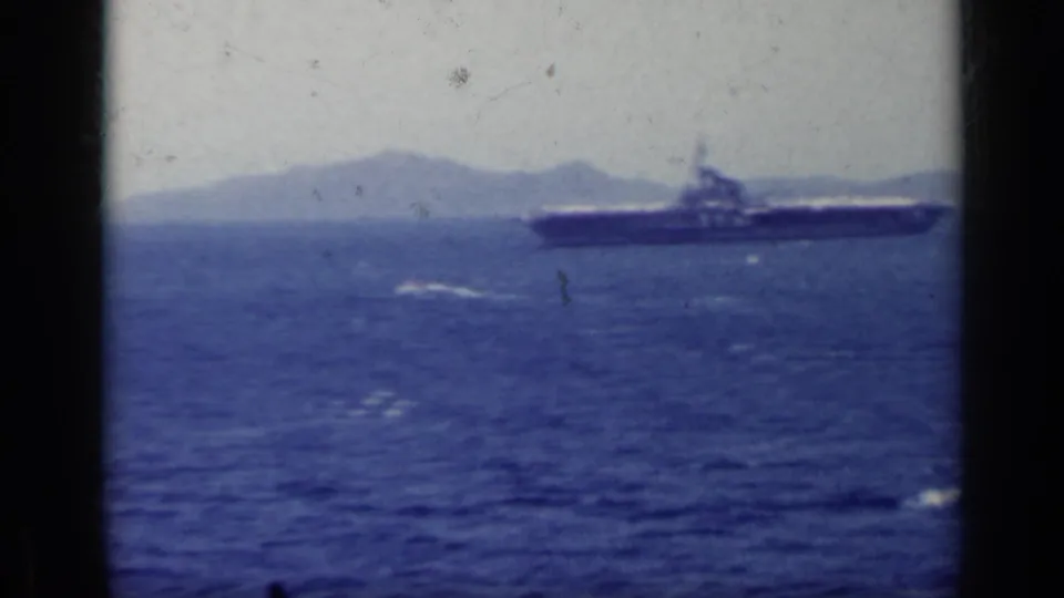 1962 Italy 8mm frame showing a naval ship crossing blue water with hazy mountains behind it.