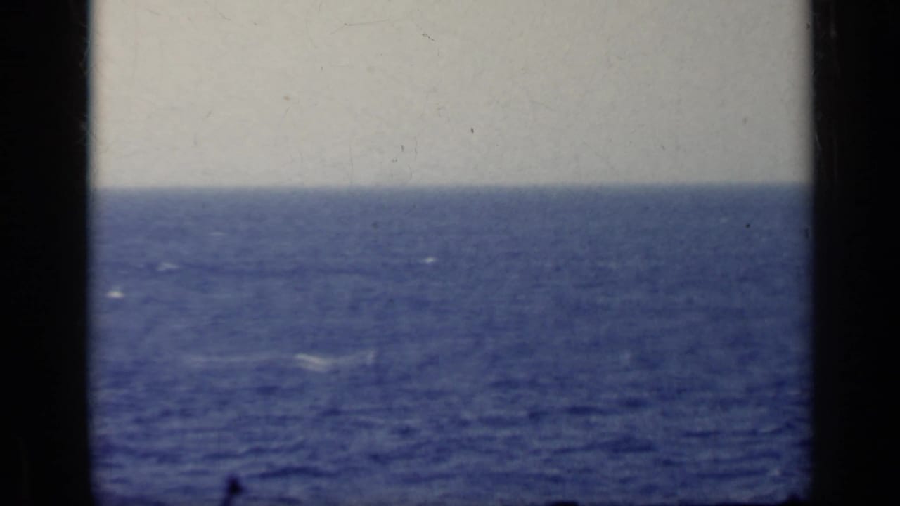 Small dark aircraft shape above a blue ocean horizon with black film borders.