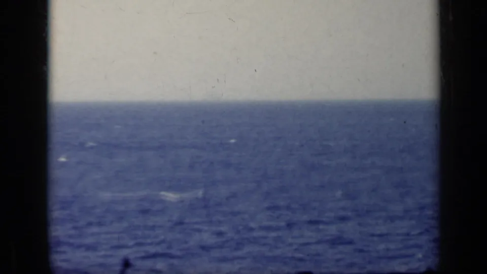 1962 Italy 8mm frame showing a distant aircraft-like silhouette over open blue water beneath a pale hazy sky.