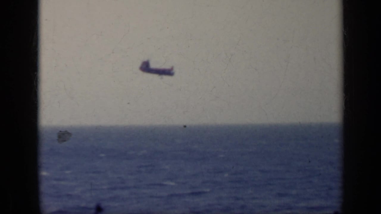 Home-movie frame from Italy in 1962 with a small dark aircraft silhouette over a broad blue sea and soft horizon.