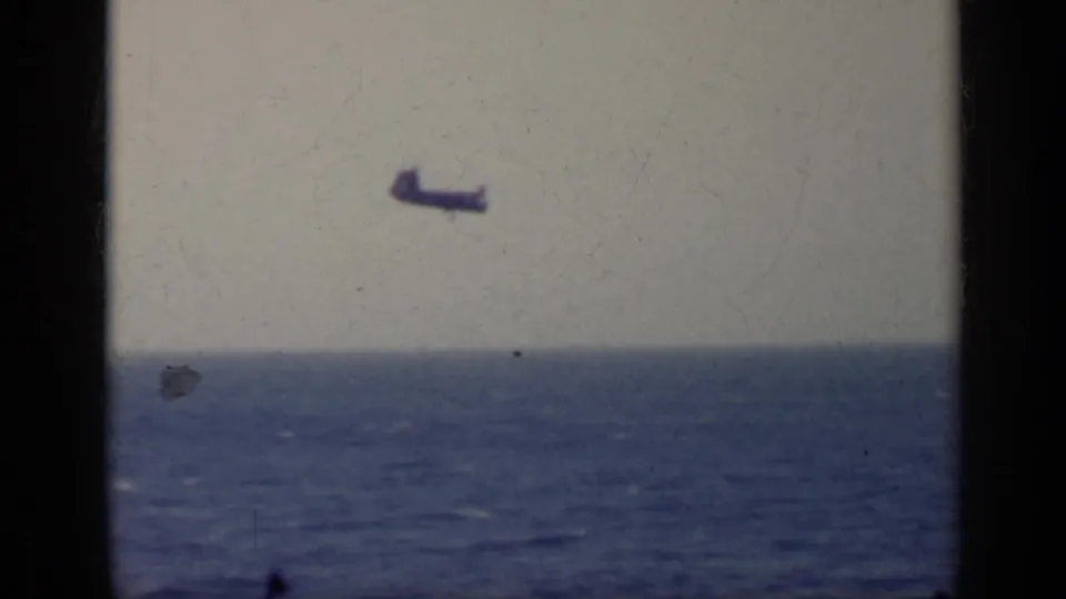 Home-movie frame from Italy in 1962 with a small dark aircraft silhouette over a broad blue sea and soft horizon.