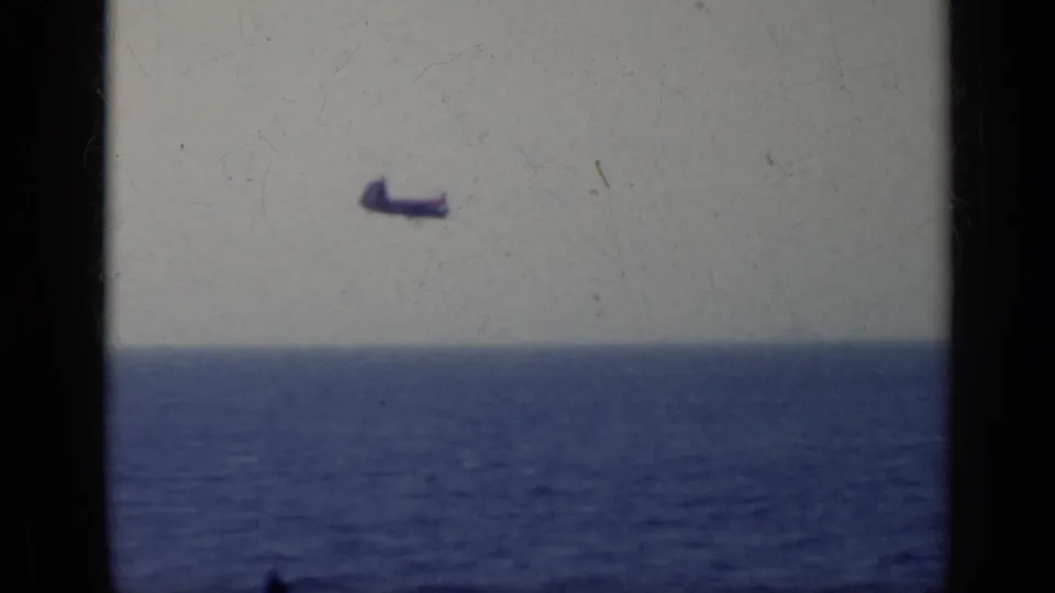 Faded 8mm frame showing a small aircraft-like mark in the sky over open water, with visible film grain and frame edge.