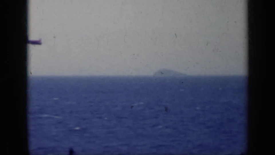 1962 Italy 8mm frame with open sea, hazy sky, a faint distant landform, and a small aircraft-like silhouette near the upper left.