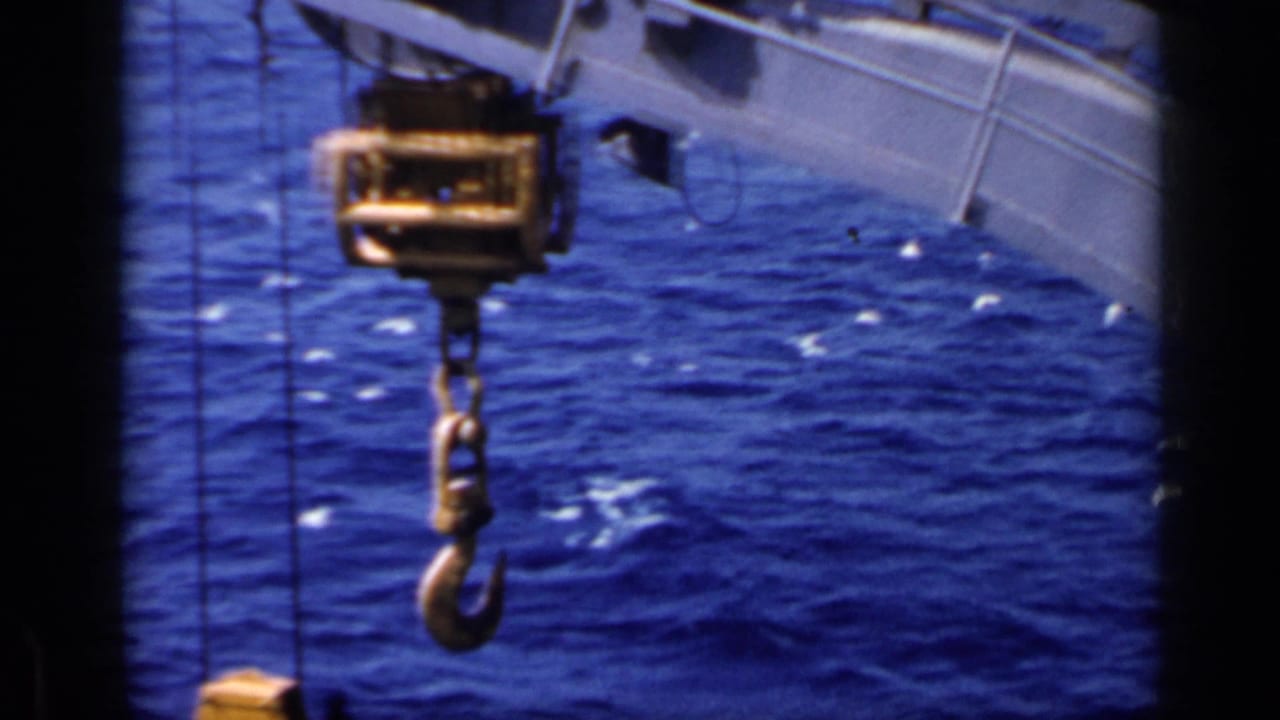 Crane hook and lifting gear hang over blue water beside the edge of a ship.