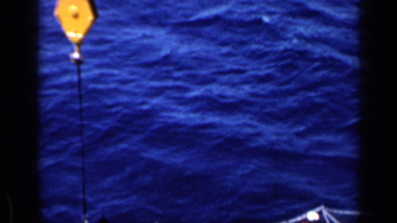 Yellow crane block hangs above rippled blue ocean with part of the ship at the edge.