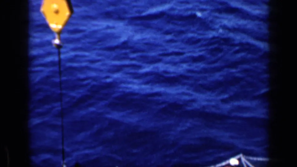 High-angle 8mm view from the 1962 Italy clip with a yellow crane block suspended over open sea from an aircraft carrier.