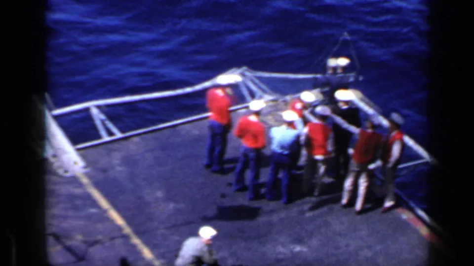 High-angle 1962 Italy 8mm frame showing uniformed crew in white caps and red or blue shirts gathered on an aircraft carrier deck.