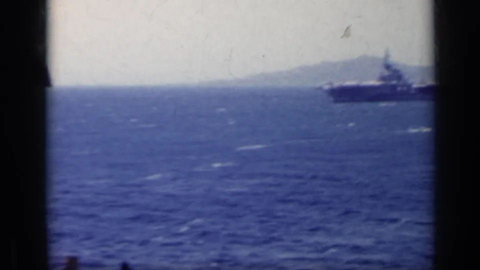 A distant naval carrier-like ship crosses blue water with a hazy Italian coastline behind it in this 1962 8mm frame.
