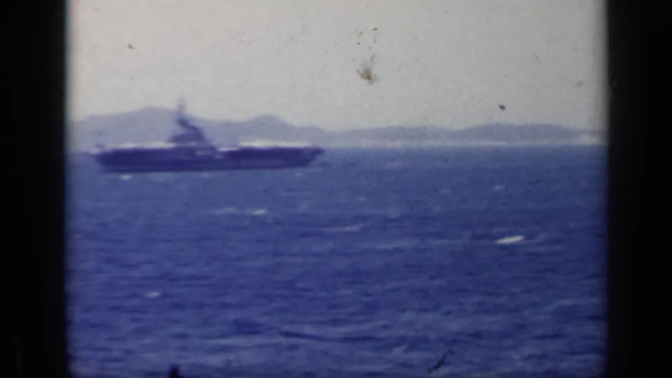 A carrier-like ship is visible on the water, with low hills and shoreline softened by haze in the background.
