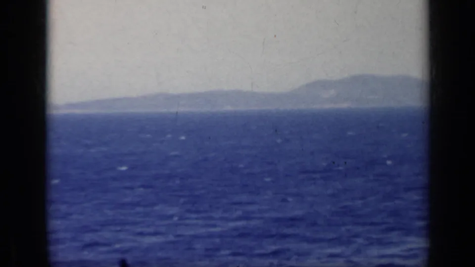1962 Italy home-movie frame showing blue sea, hazy coastal mountains, and dark window or film-gate framing.