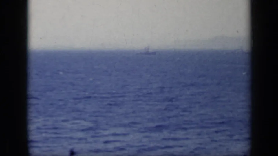 1962 8mm Italy frame showing blue sea, pale sky, dark foreground bars, and a faint distant vessel or shoreline shape near the horizon.