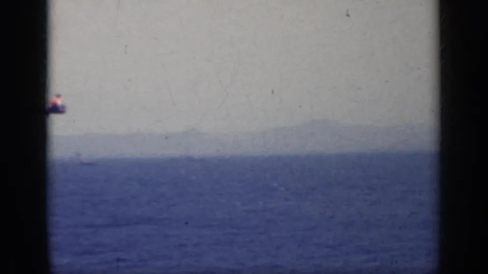 A small red and white aircraft begins to pass into a 1962 Italy sea view, with hazy landforms beyond the water.