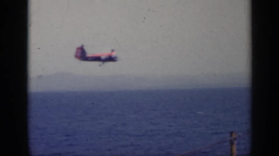 Red and white aircraft crossing above the blue sea in 1962 Italy footage, with a soft hazy coastline in the distance.