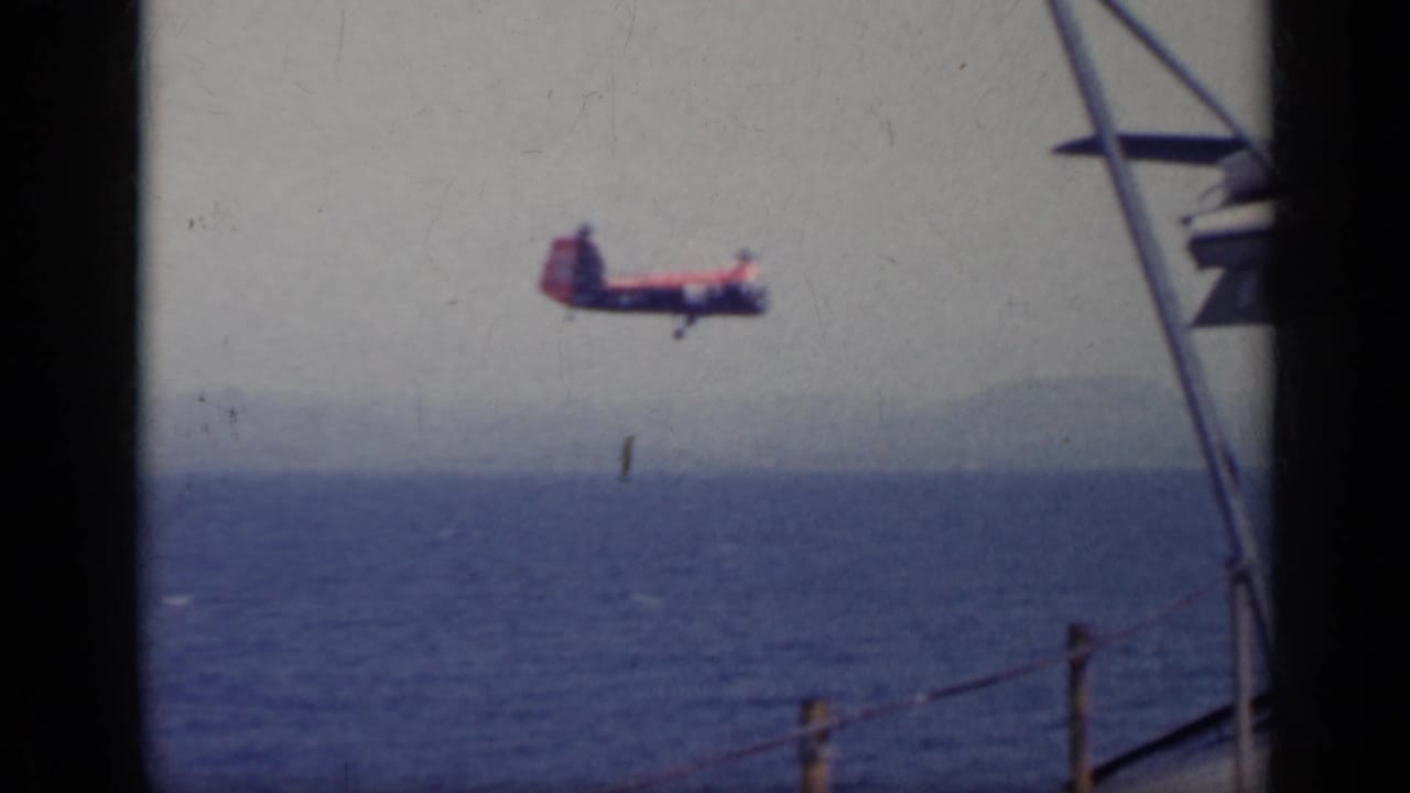 Red and white helicopter flying over the sea near a ship rail.