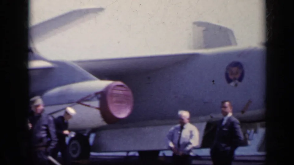 People stand near a large military aircraft on a carrier deck, with fuselage markings visible in 1962 footage.