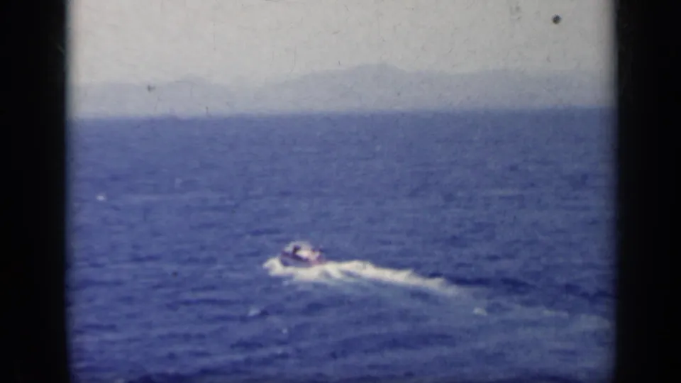 The small boat continues across open water, with haze on the horizon and 8mm film wear visible.