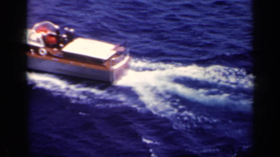 The speedboat passes partly out of frame, its white wake spreading across saturated blue water in 8mm footage.