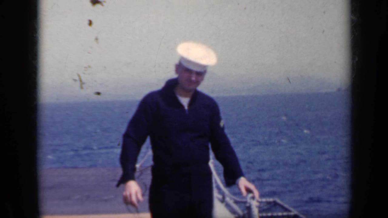 Sailor in dark uniform and white cap standing on a ship deck with the ocean behind him.