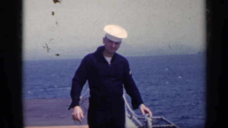 1962 Italy 8mm frame of a sailor in a dark naval uniform and white cap on an open ship deck, with blue sea and pale sky behind.