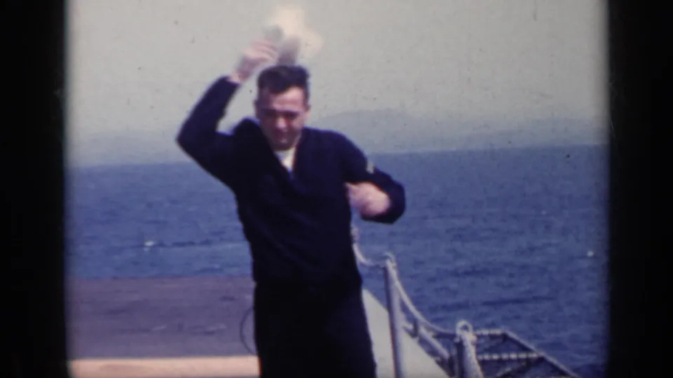 1962 Italy 8mm frame showing a sailor lifting or losing a white dixie cup cap in strong wind on a ship deck at sea.