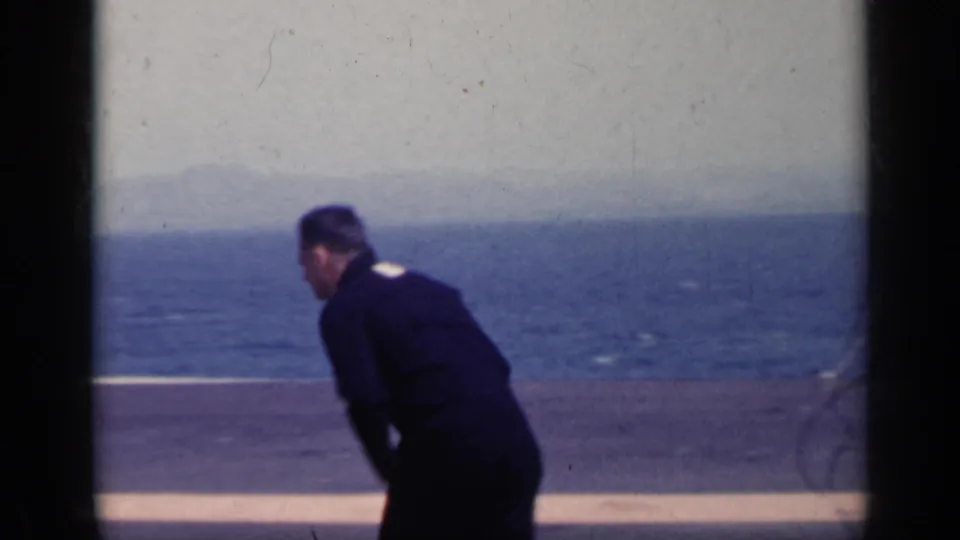 1962 Italy 8mm frame of a sailor leaning down on deck after the windblown cap moment, with open sea visible beyond the rail.