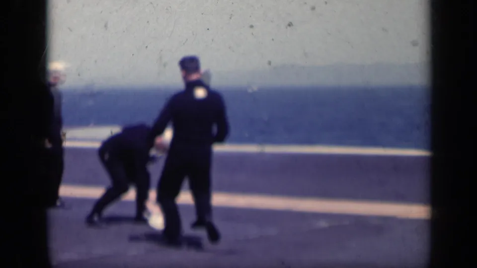 1962 Italy 8mm frame showing sailors on an open ship deck, one bending near the ground while others stand nearby against the ocean backdrop.