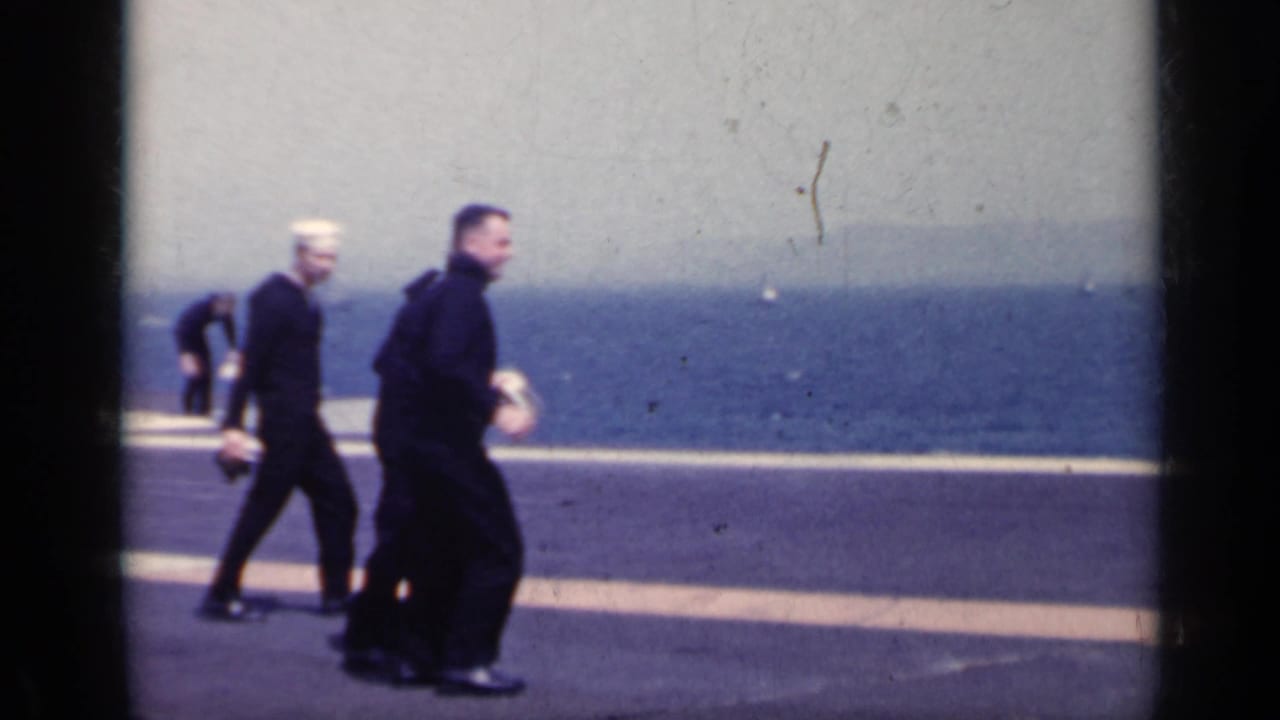 Sailors in dark uniforms and white hats walk across a carrier deck with ocean behind them.