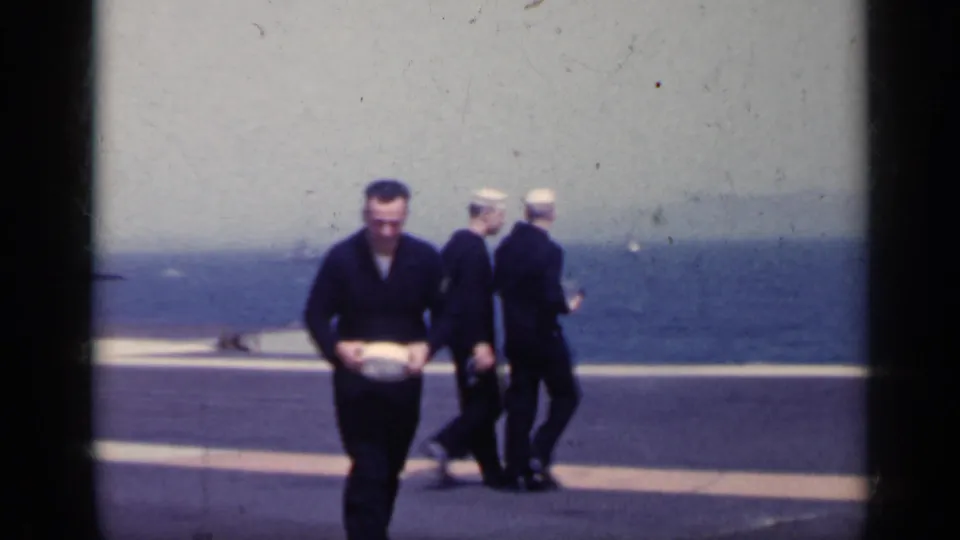 A U.S. Navy sailor in dark uniform and white hat approaches the camera as other sailors move across the carrier deck at sea.
