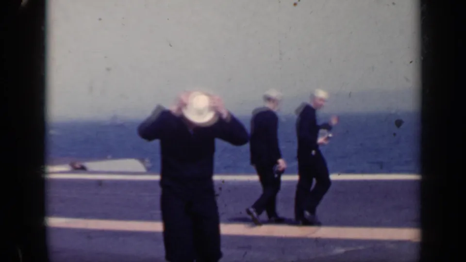 1962 carrier-deck frame showing a sailor adjusting his white hat in the foreground as two sailors walk behind him against the sea.