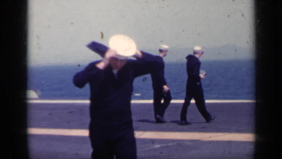 Close foreground view of a U.S. Navy sailor adjusting his white hat on a carrier deck, with two sailors and the sea behind him.