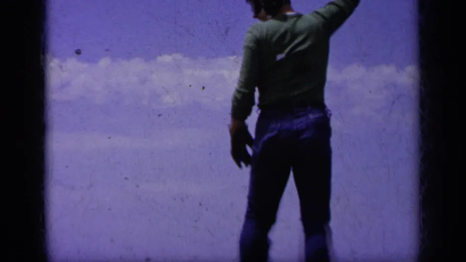 A green-shirted deck signaler, seen from behind in the 1963 Greece footage, raises an arm against the sky during carrier operations.