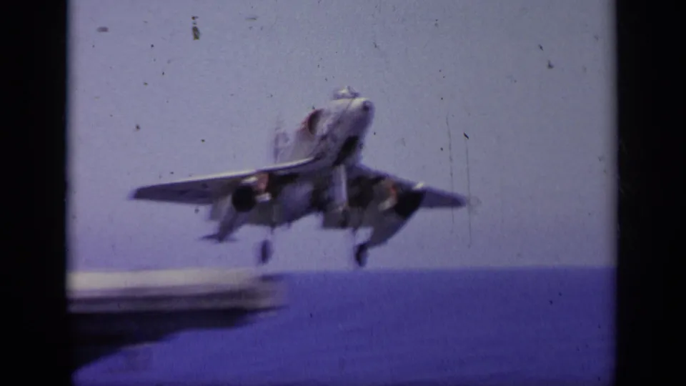 1963 Greece home-movie frame of a jet aircraft airborne off a carrier deck, seen against sea and sky.