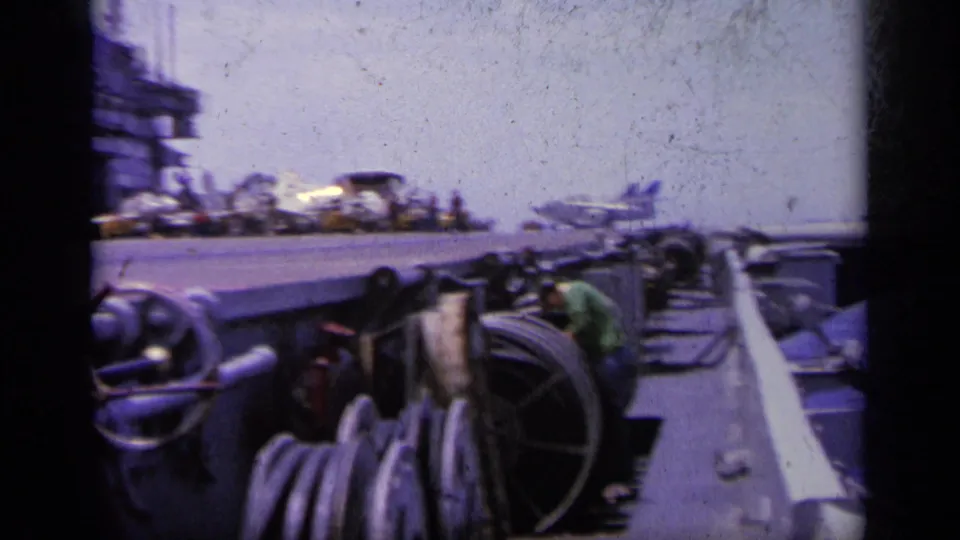 1963 Greece home-movie frame of carrier deck equipment, railings, crew area, and an aircraft near the deck edge.