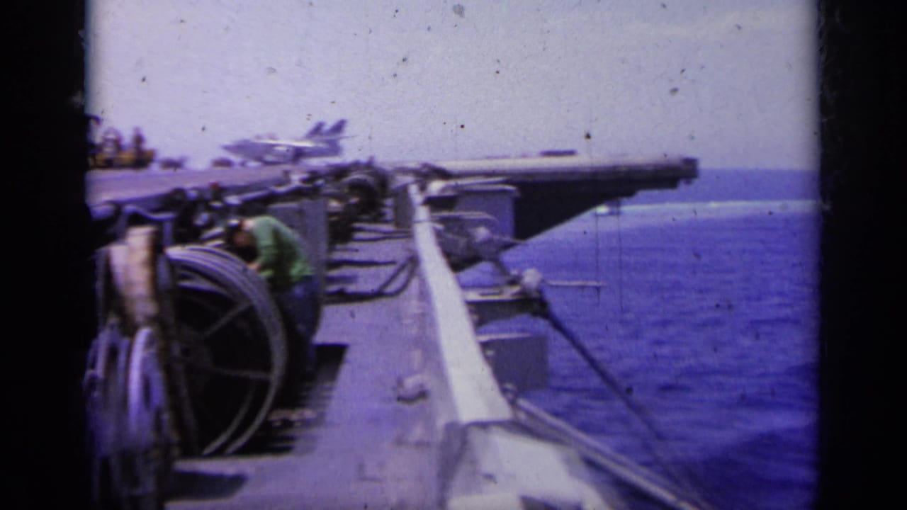 Aircraft carrier deck edge with cable reels, crew, sea, and a jet in the distance.