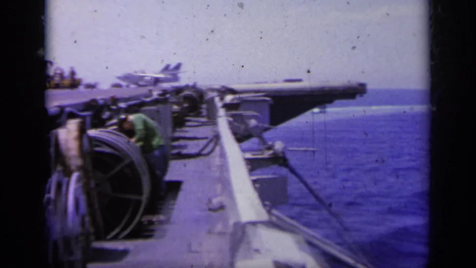1963 8mm view from an aircraft carrier near Greece, showing deck equipment, crew figures, ocean, and a distant military jet.