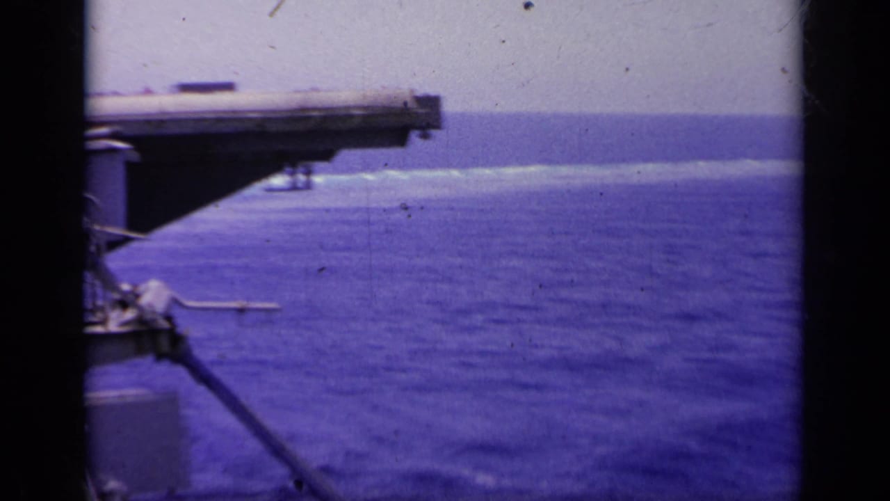Carrier deck overhang and open ocean with a small aircraft shape near the water.