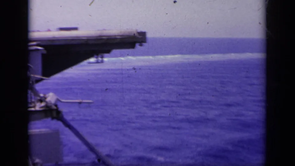 Handheld 1963 carrier-side view near Greece, with deck structure at left and open sea stretching to the horizon.