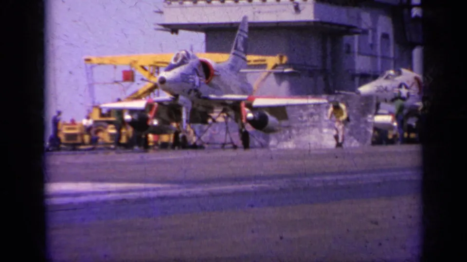 1963 8mm view of a parked military jet on an aircraft carrier deck near Greece, with crew and deck equipment visible.