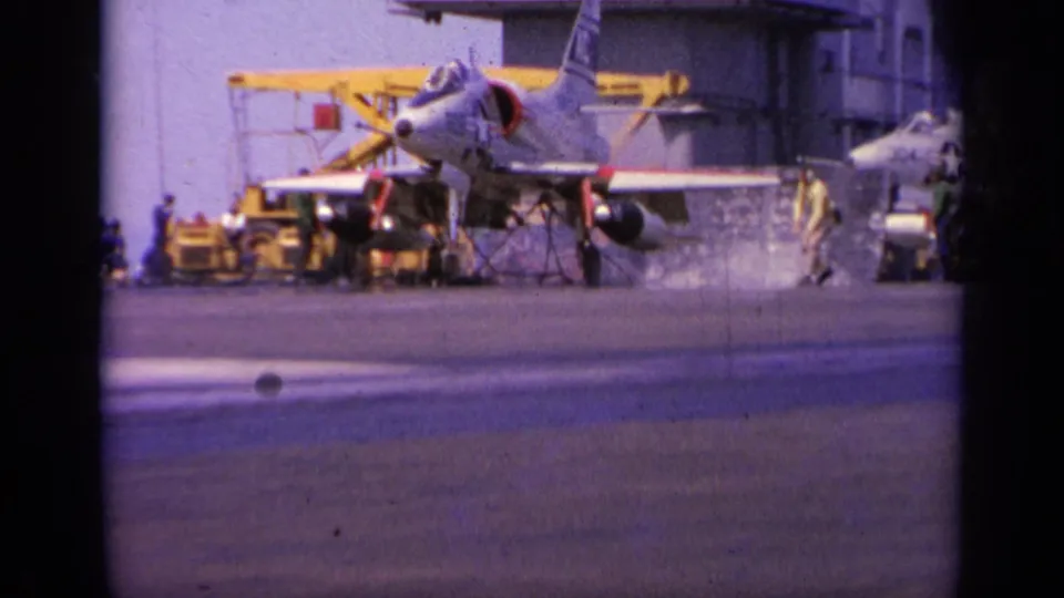 1963 Greece 8mm frame showing a military jet on a carrier deck, with crew, yellow deck equipment, and the island structure behind it.