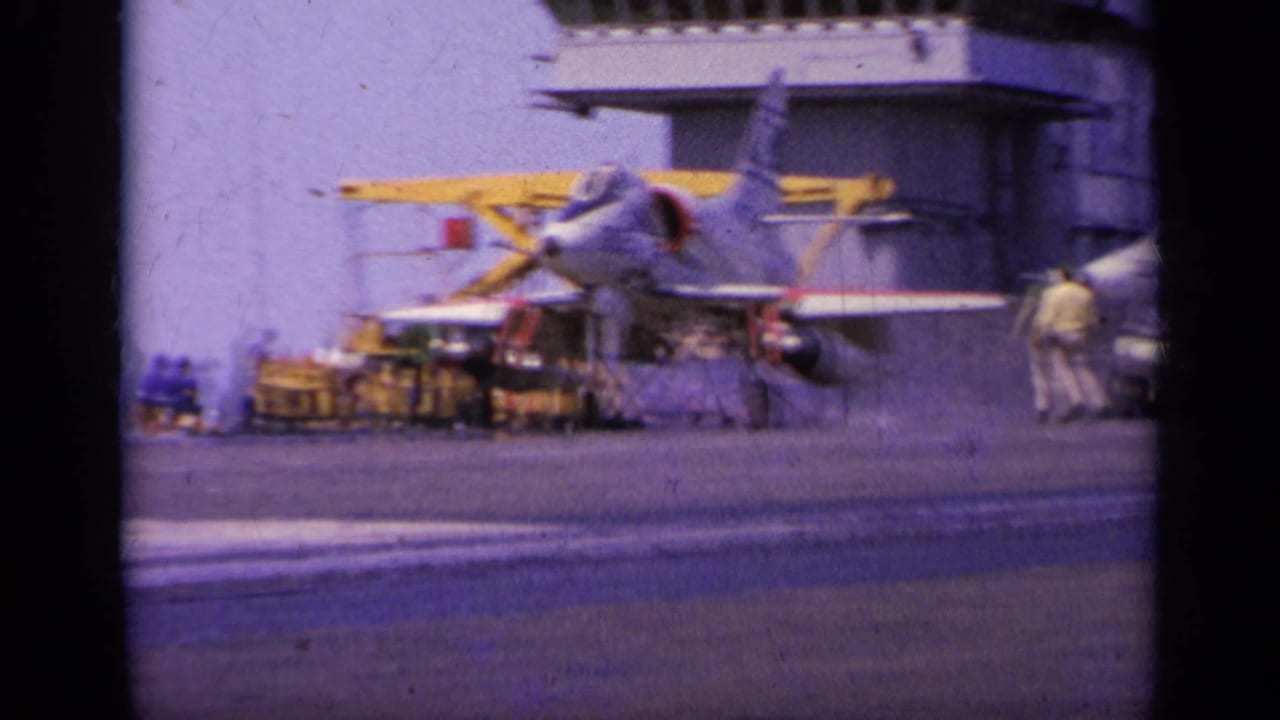 Military jet on carrier deck with crew member and deck equipment near the aircraft.