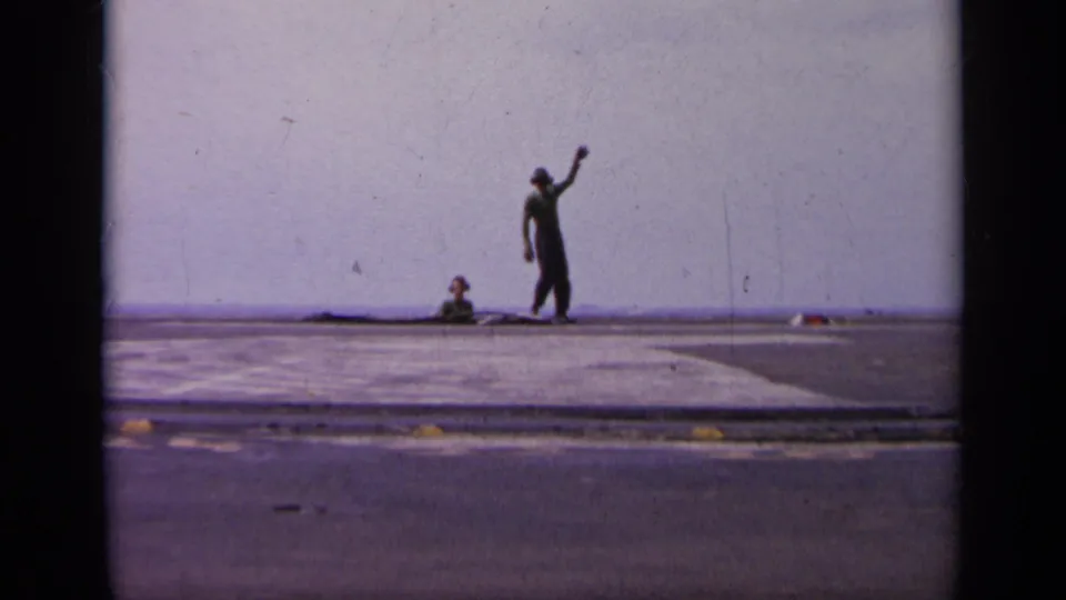 A deck crew member signals on the carrier runway, with the open sea horizon and deck surface visible in the 1963 Greece footage.