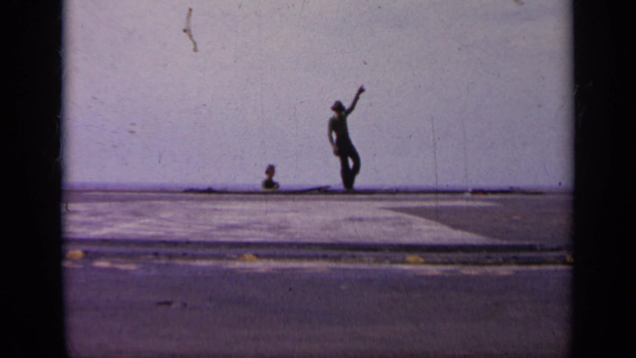 Distant crew member signals on an open aircraft-carrier deck near a hazy horizon.