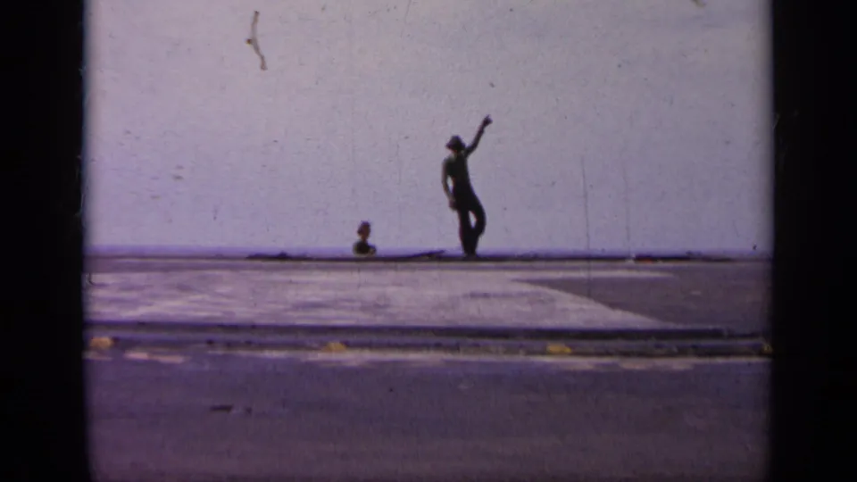 1963 Greece 8mm frame showing a distant crew signaler on a broad aircraft-carrier deck with sea and sky beyond.